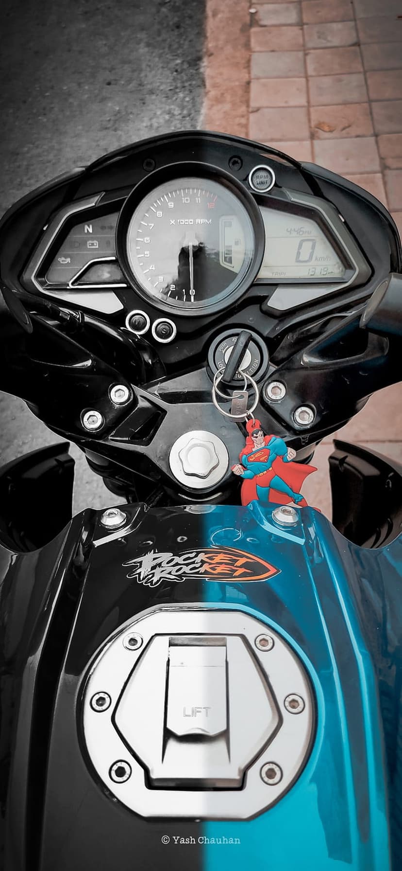 Motorcycle speedometer and handlebars with a Superman keychain and Pocket Rocket decal on the fuel tank.