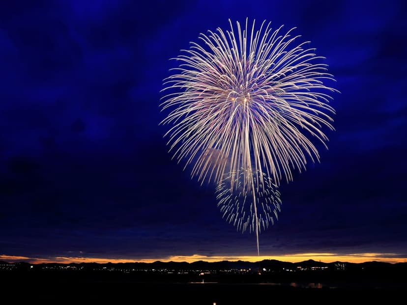 Gold and white fireworks explode over a dark landscape silhouette and a hazy orange horizon in a night sky.