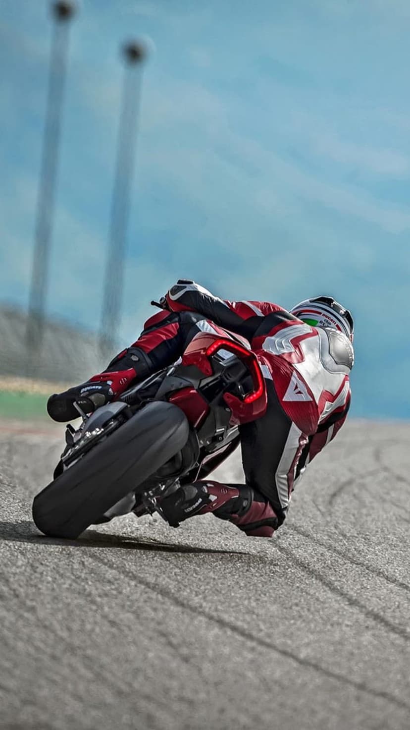 Motorcyclist in red and white gear leans low into a turn on a grey asphalt race track under a blue sky.