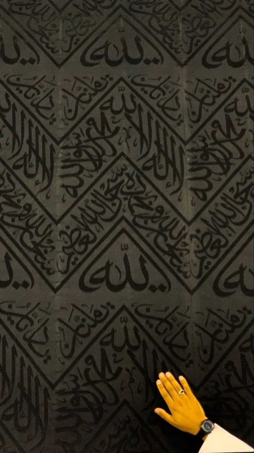 Gold Arabic calligraphy on black silk Kiswah fabric as a hand wearing a ring touches the sacred textile.