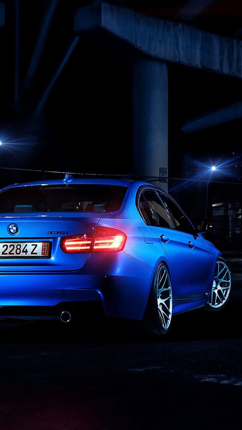 Blue BMW 335i sedan with glowing red taillights parked under a concrete bridge at night with an urban backdrop.