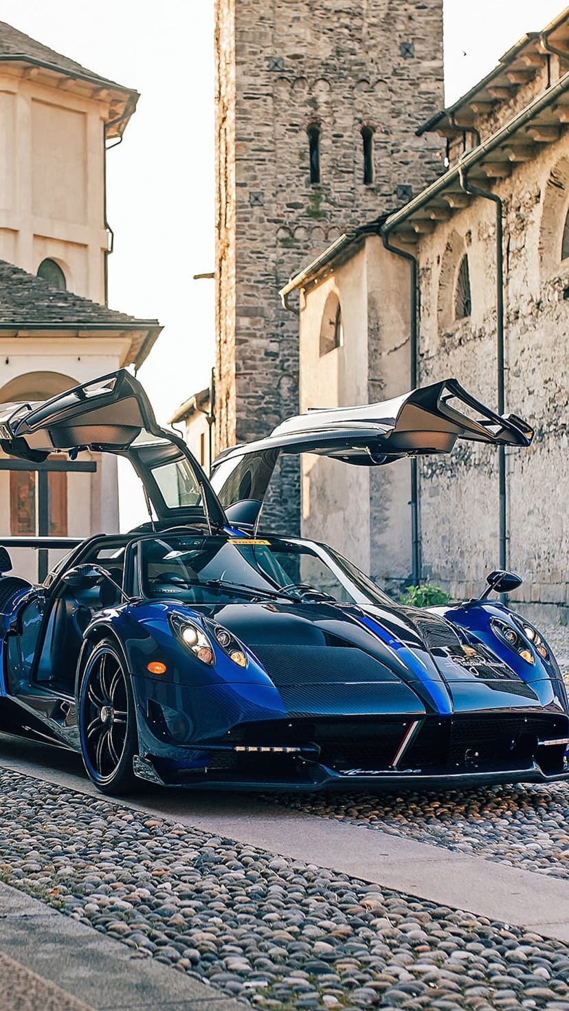 Blue Pagani Huayra Roadster with open butterfly doors parked on an Italian cobblestone street during sunset.