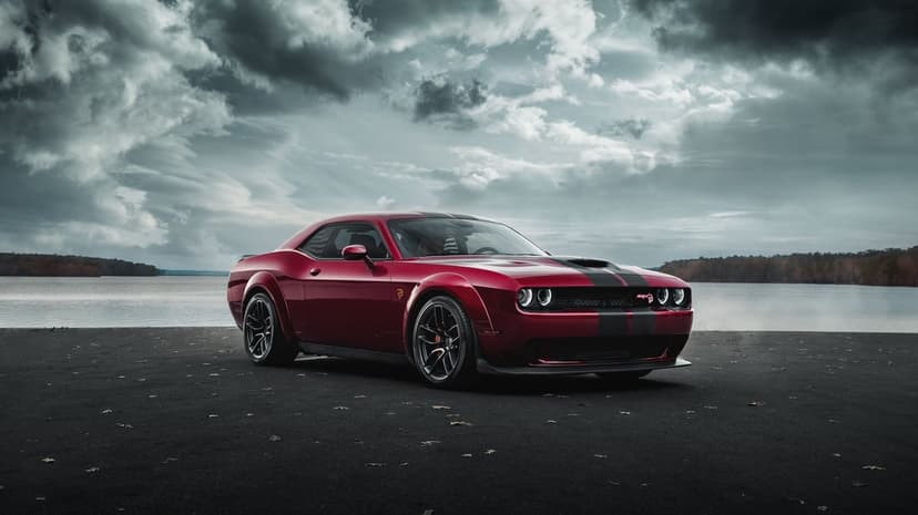 Red Dodge Challenger SRT Hellcat Widebody with black stripes parked by a lake under a dark stormy sky.