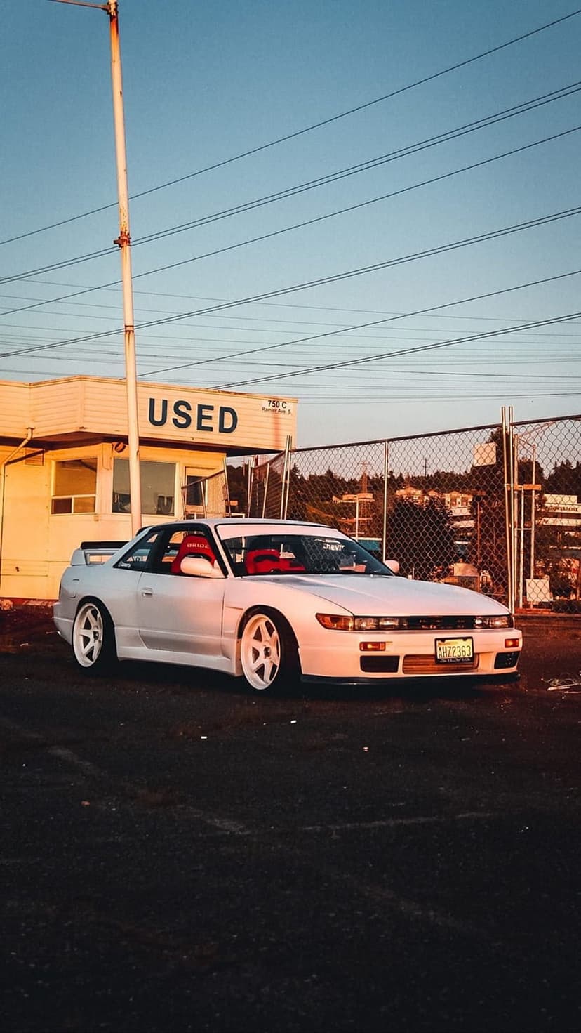 White Nissan S13 coupe with red bucket seats parked by a chain-link fence during a warm golden hour sunset.