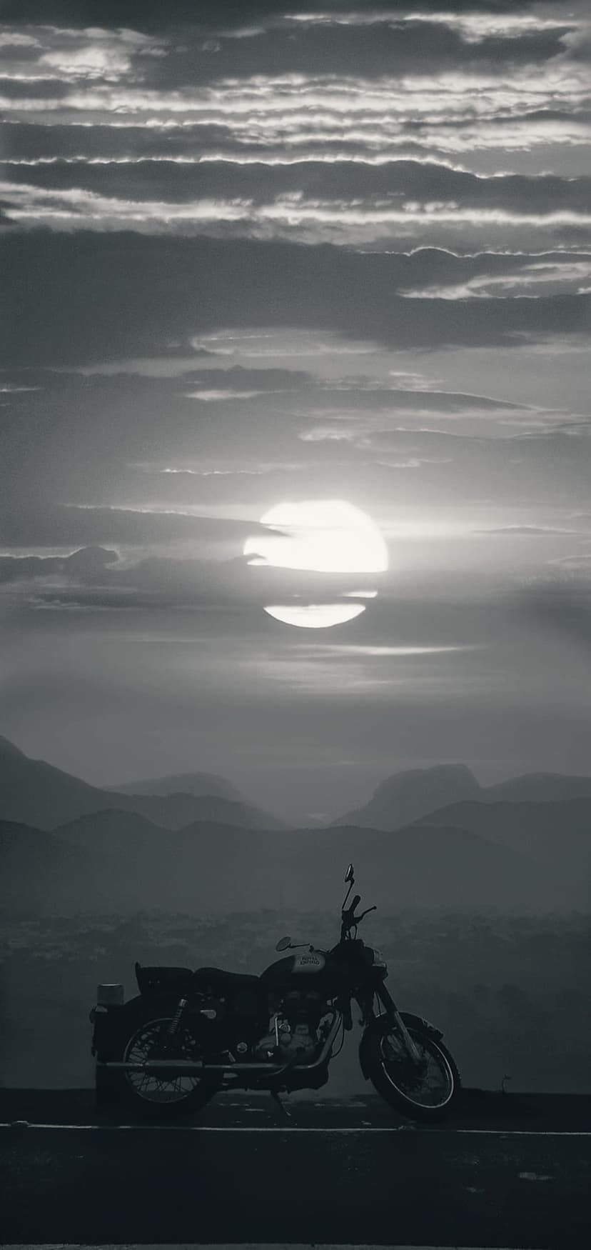 Motorcycle silhouette on a mountain ridge against a cloudy sunset with misty hills fading into the background.