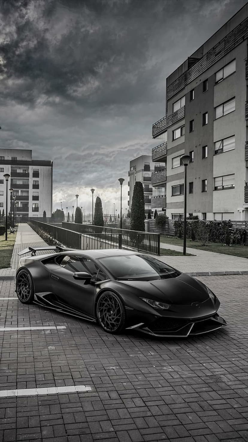 Black Lamborghini Huracan parked on cobblestone pavement against modern urban architecture and cloudy skies.
