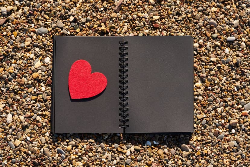 Red felt heart lies on a black notebook over a bed of rounded, colorful pebbles in a vertical composition.