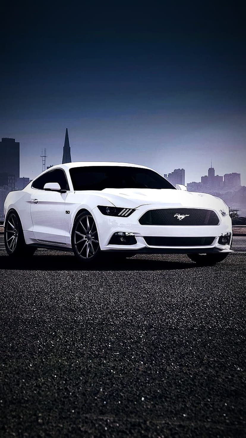 White Ford Mustang coupe parked on dark textured asphalt with a blurred cityscape and deep blue sky behind it.