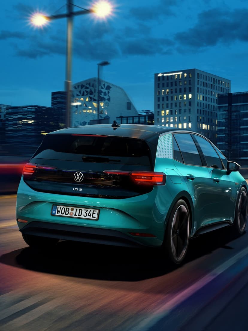 Teal Volkswagen ID.3 electric car driving on an urban road at night past glowing buildings with motion blur.