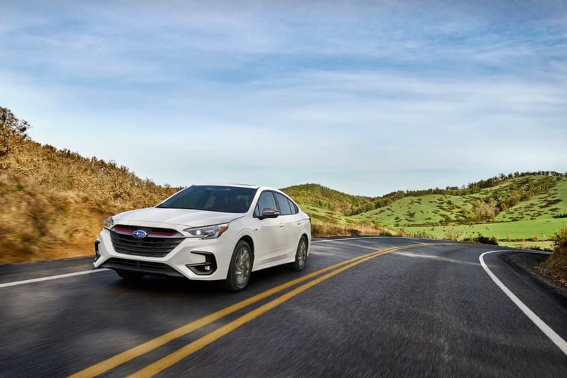 White Subaru sedan driving on a curved road with yellow lines through green rolling hills under a blue sky.