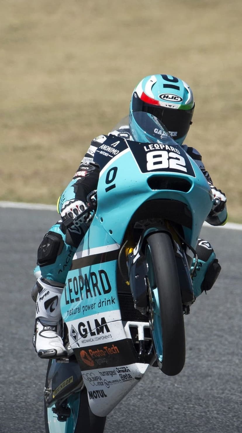 Motorcycle racer in a turquoise suit pulls a wheelie on a branded sportbike with the front wheel in the air.