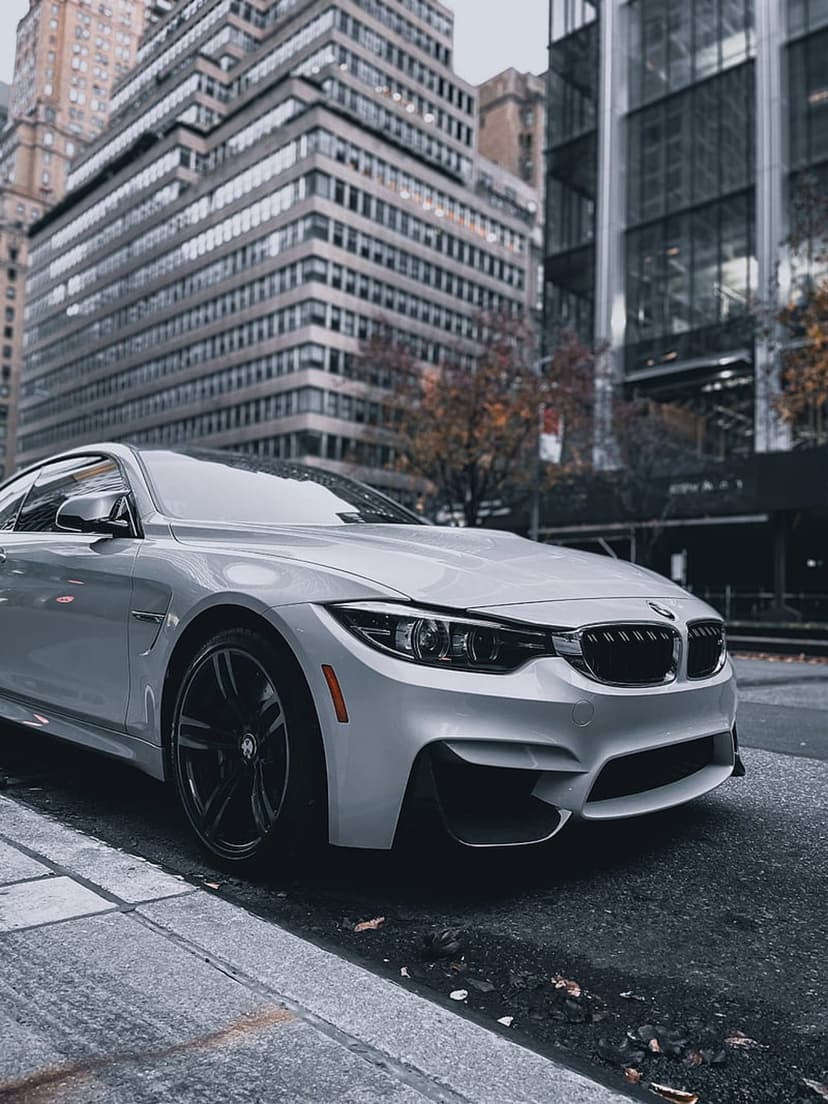 White BMW M4 coupe parked on a wet asphalt street framed by tall skyscrapers and fallen autumn leaves at a low angle.