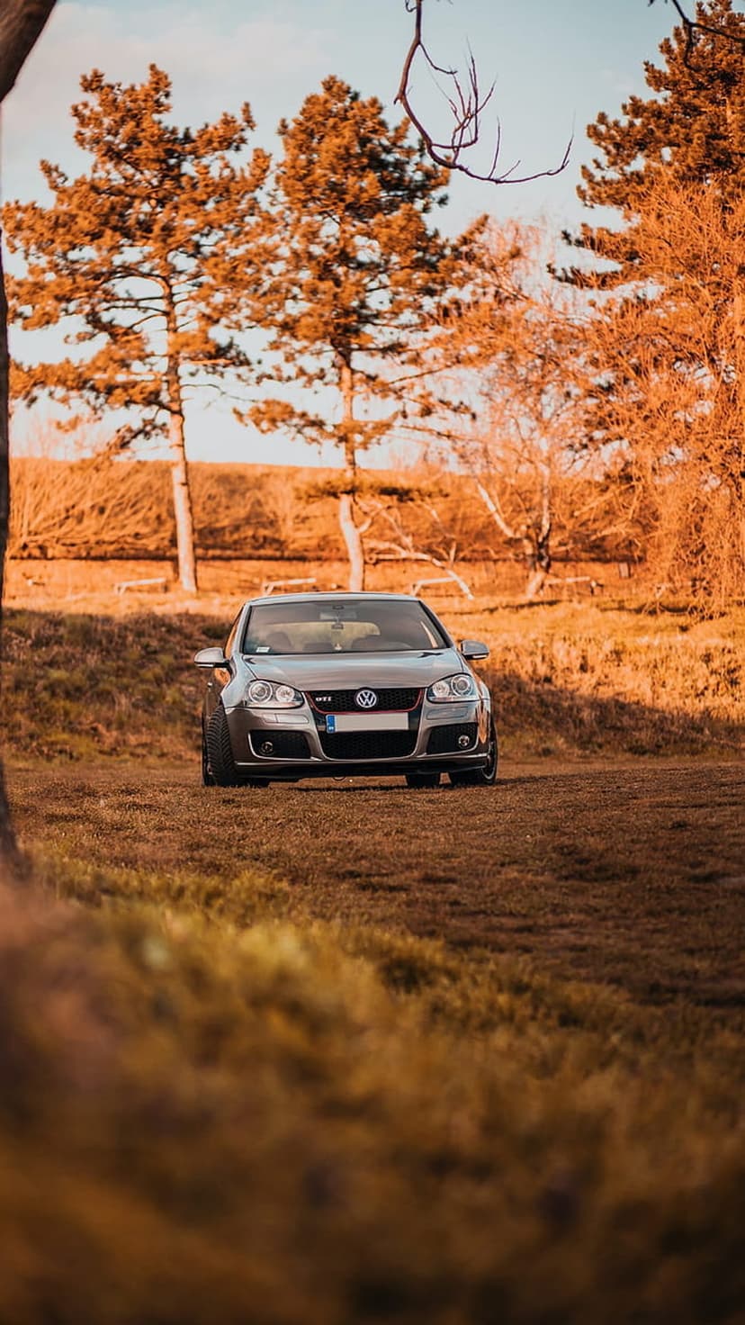 Silver Volkswagen GTI parked in a grassy field surrounded by vibrant orange and yellow autumn trees at low angle.