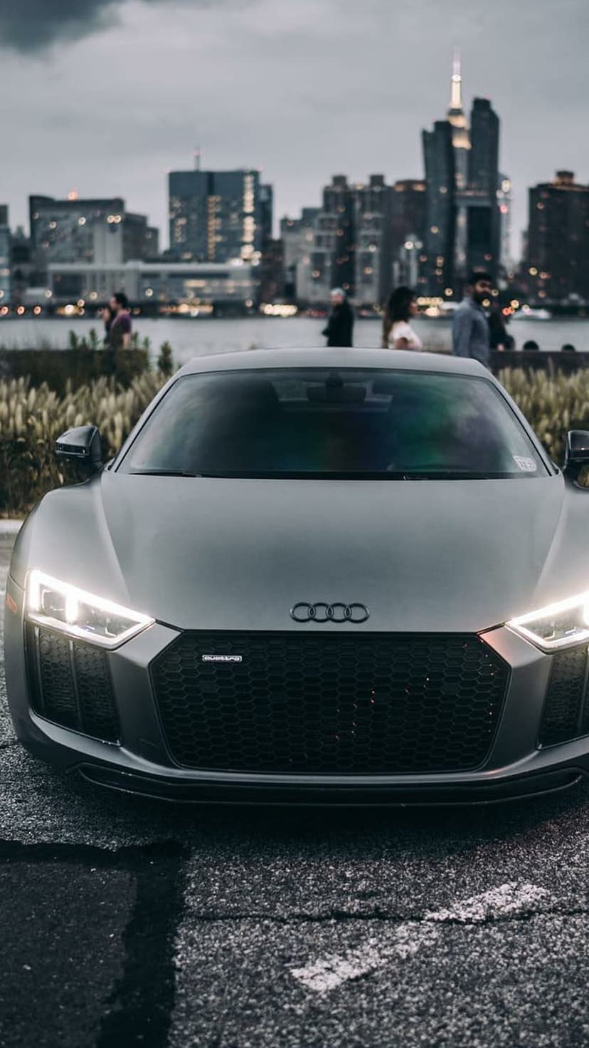 Audi R8 front grille and glowing headlights parked on asphalt with a blurred night city skyline in the distance.