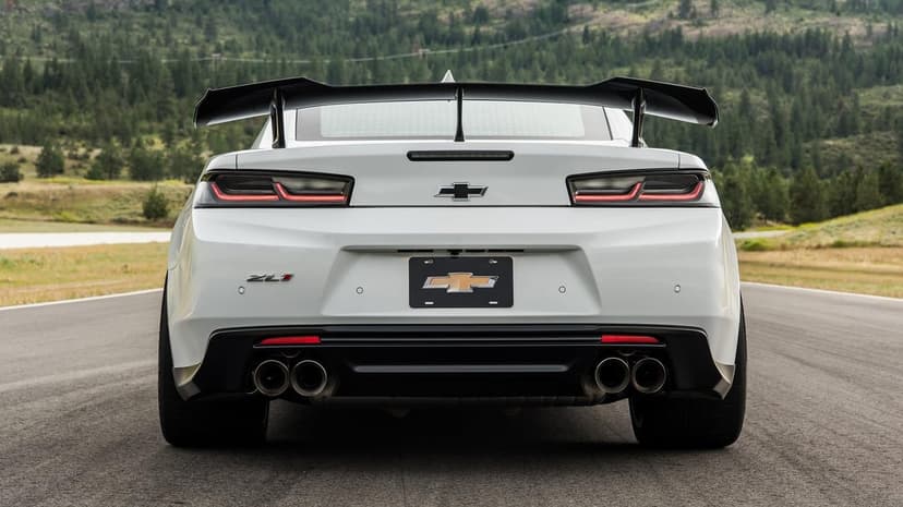 Rear view of a white Camaro ZL1 with a carbon fiber wing on an asphalt track against a forest backdrop.