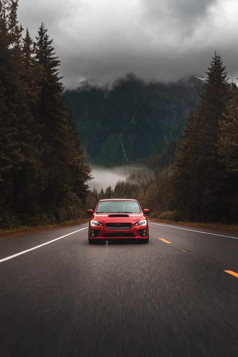 Red Subaru WRX driving on a wet asphalt road through a misty pine forest with mountains and a distant waterfall.