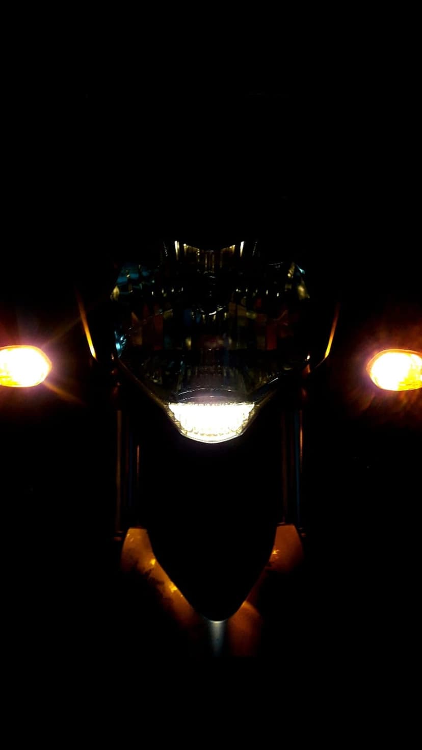 Motorcycle headlight and bright amber turn signals glow intensely against a dark, shadowy urban night background.
