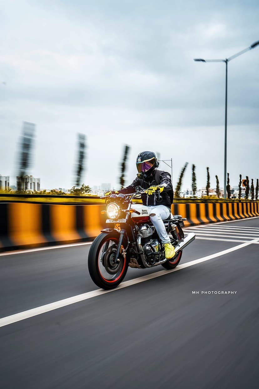 Red and white motorcycle rider in a black jacket and yellow shoes traveling on a blurred highway under gray clouds.