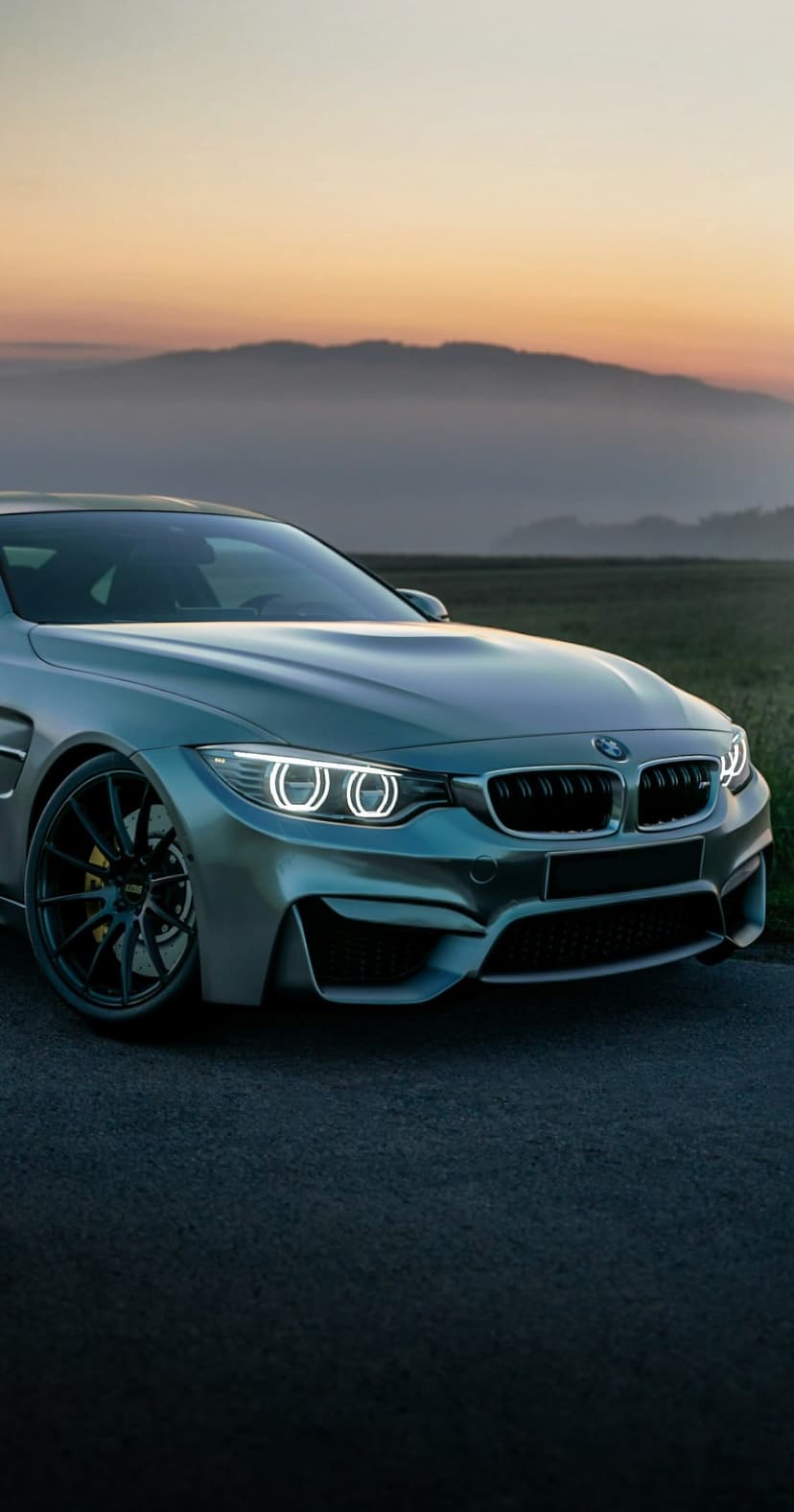 Silver BMW M4 Coupe with glowing headlights on an open road at sunset against a misty mountain backdrop.