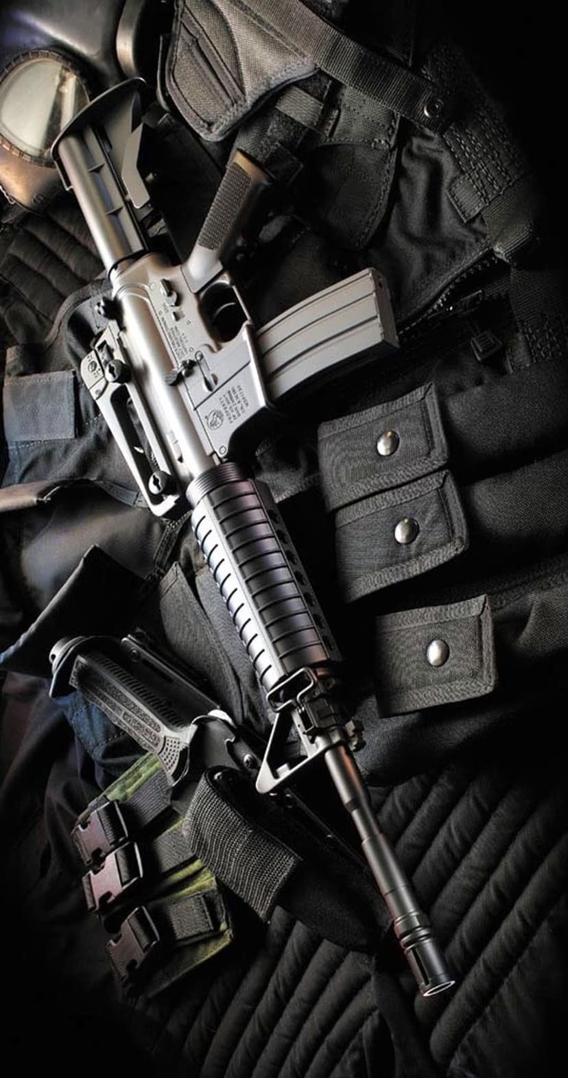 AR-style rifle and a holstered handgun positioned on a black tactical vest under dramatic low-key lighting.