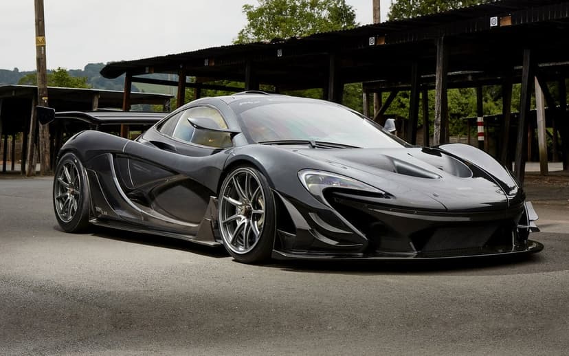 McLaren P1 LM in dark gray parked on asphalt under a wooden shelter with trees in the background.