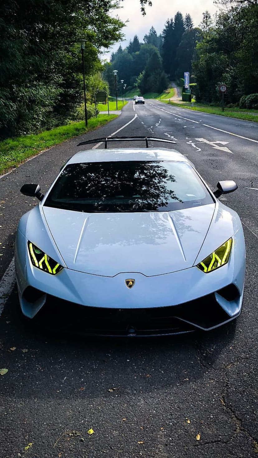 White Lamborghini Huracan with yellow headlights on a wet forest road surrounded by dense green trees and foliage.