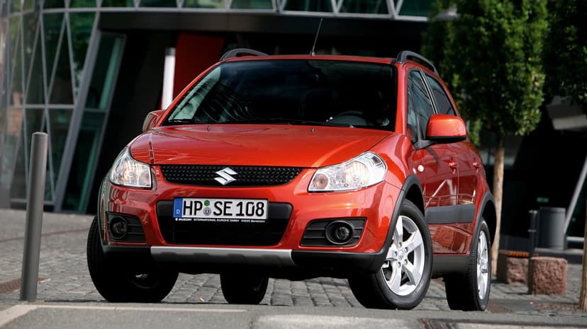 Red Suzuki SX4 compact SUV parked on a cobblestone city street in front of a modern glass building.