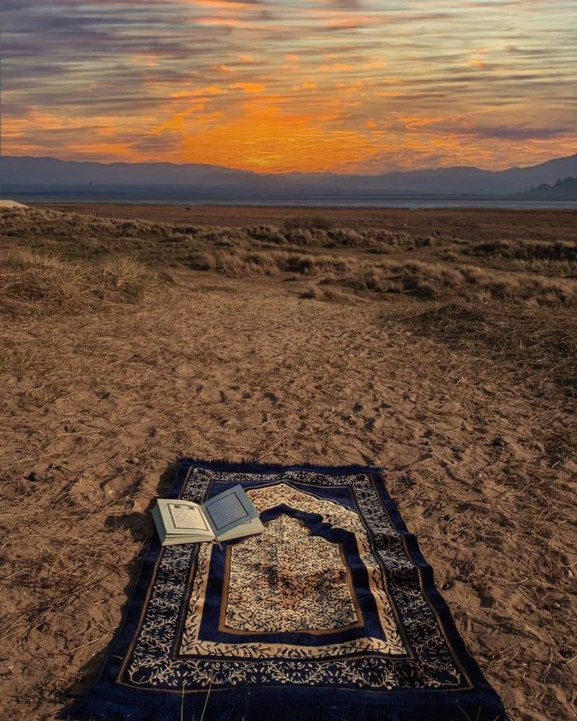 Open Quran on a prayer rug at a sandy beach beneath a vibrant purple sunset with dunes and distant mountain peaks.