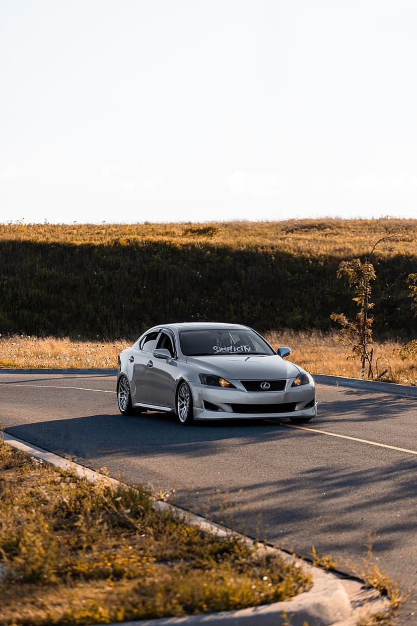 Silver Lexus IS sedan driving on a paved rural road past golden grassy hills during a bright sunny day.