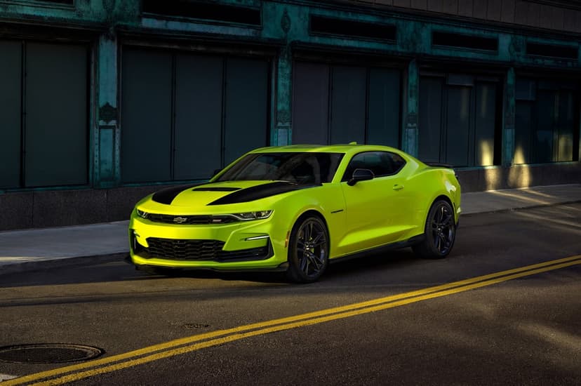 Neon yellow Chevrolet Camaro with black hood stripes parked on an asphalt street in front of a teal building.