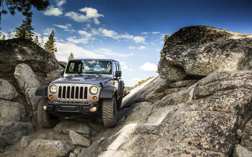 Silver Jeep Wrangler Rubicon off-roading through a narrow rocky gap under a blue sky with white clouds.