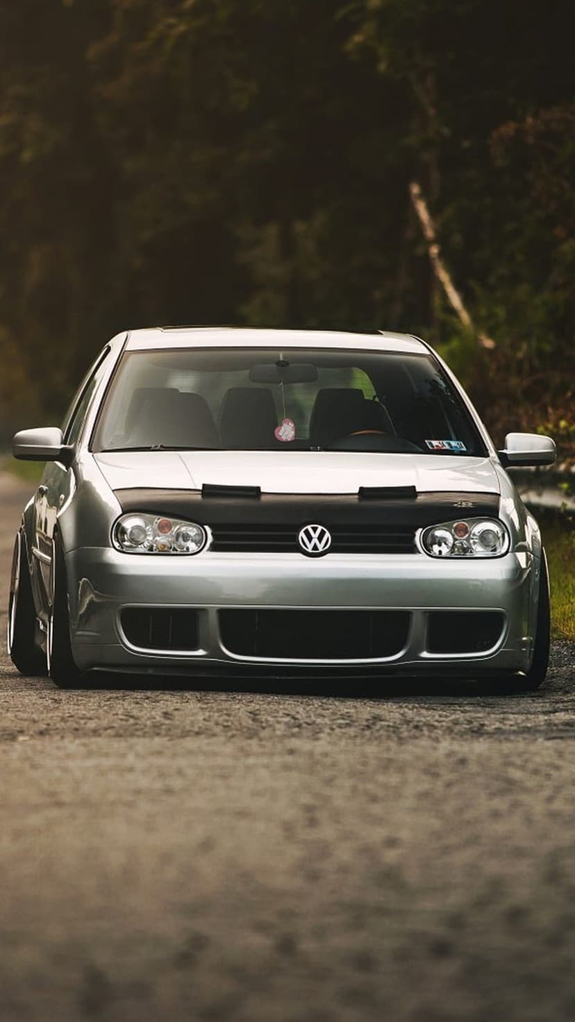 Silver VW Golf GTI with a black hood bra and custom wheels parked on a gravel road surrounded by tall trees.