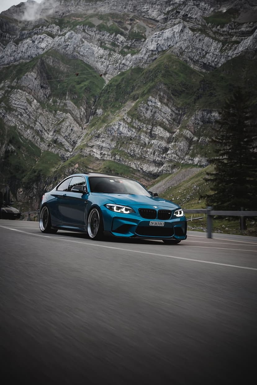 Blue BMW M2 sport coupe driving on a winding mountain road against a backdrop of jagged, rocky alpine peaks.