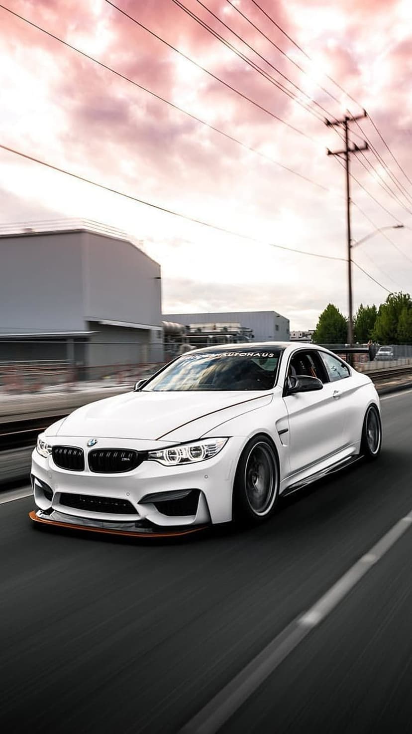 White BMW M4 coupe speeds down a road during a pink sunset with industrial buildings in the background.