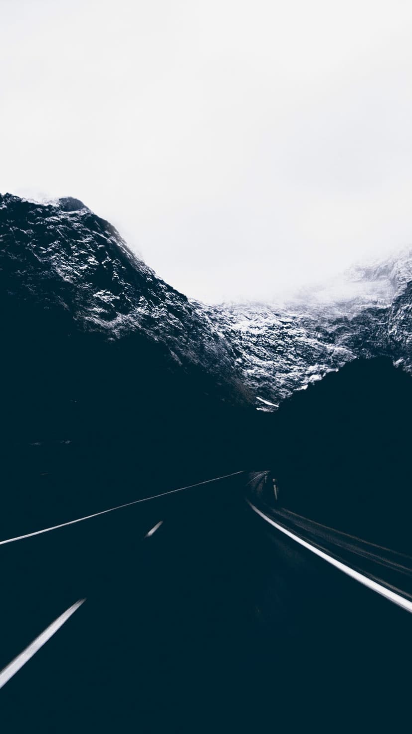Dark asphalt road winding through steep, snow-capped mountain peaks under a pale, overcast winter sky.