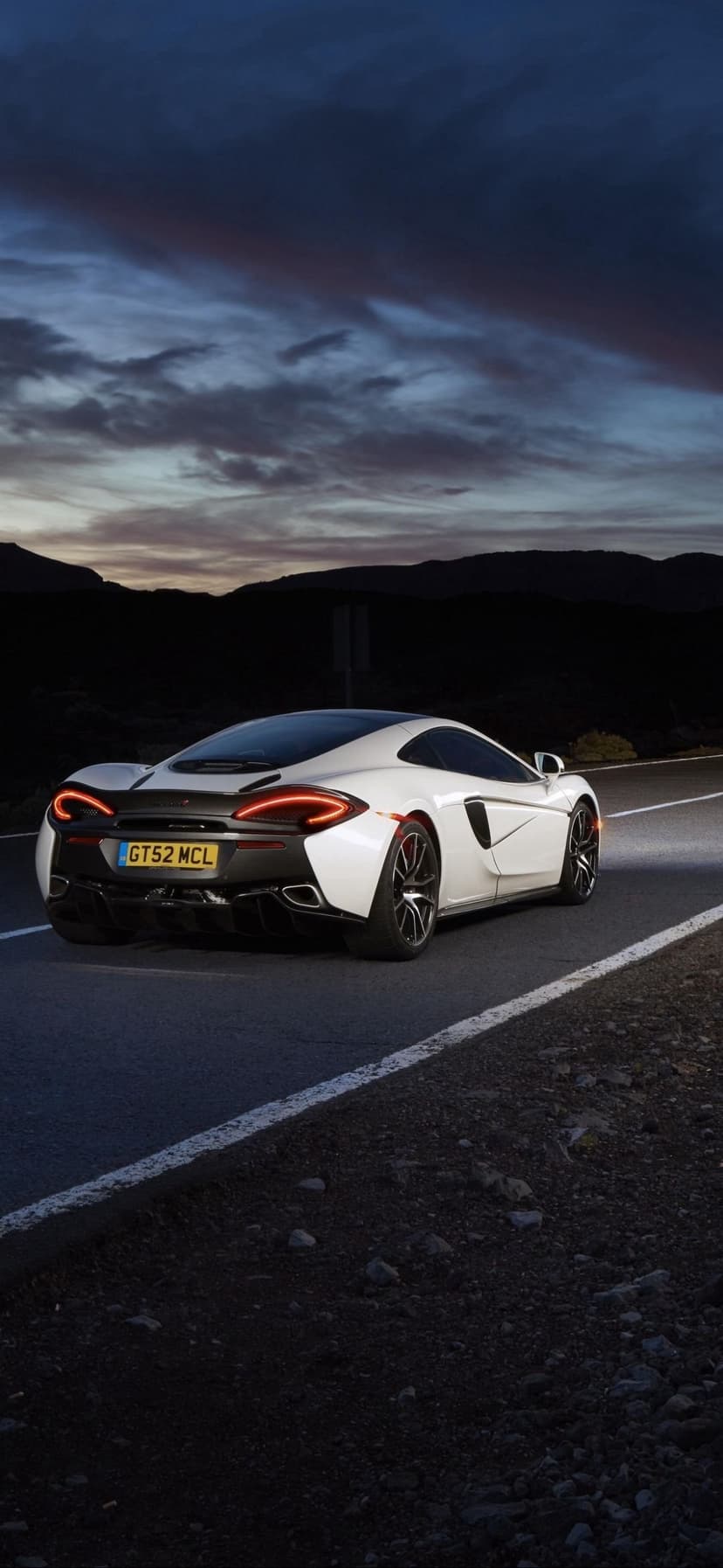 White McLaren 570S rear view with glowing taillights on a road against a purple sunset and mountain silhouette.