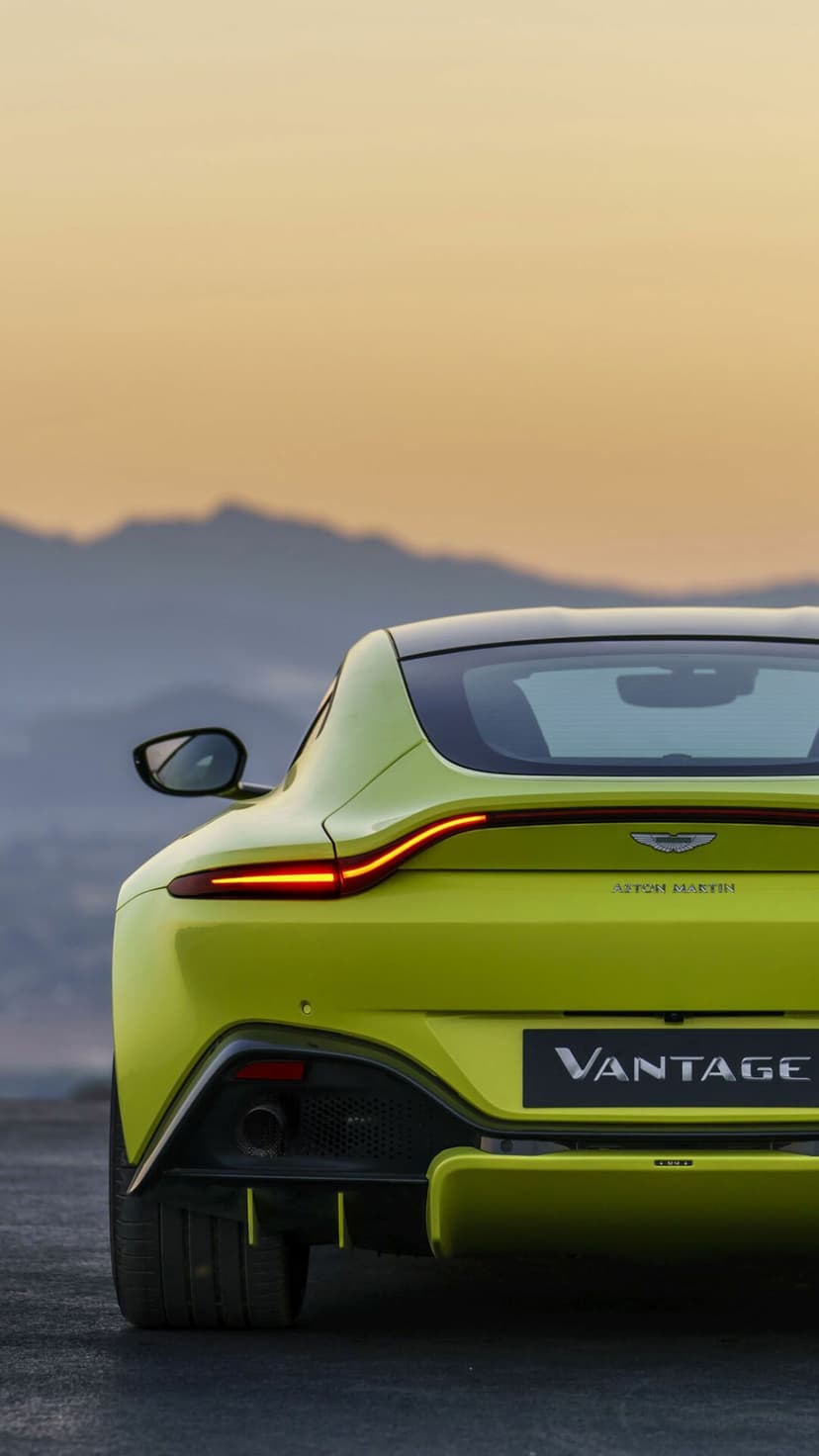 Rear of a lime green Aston Martin Vantage with glowing taillights against a soft mountain sunset backdrop.