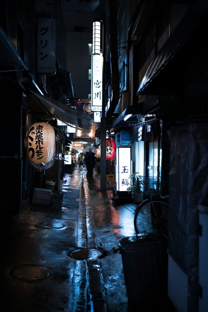 Glowing neon signs reflect on a rainy Japanese alleyway with wet cobblestones and distant figures under umbrellas.