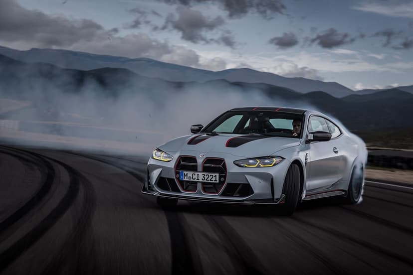 Silver BMW M4 CSL sports car drifting on a dark track with thick white smoke against a mountain backdrop.