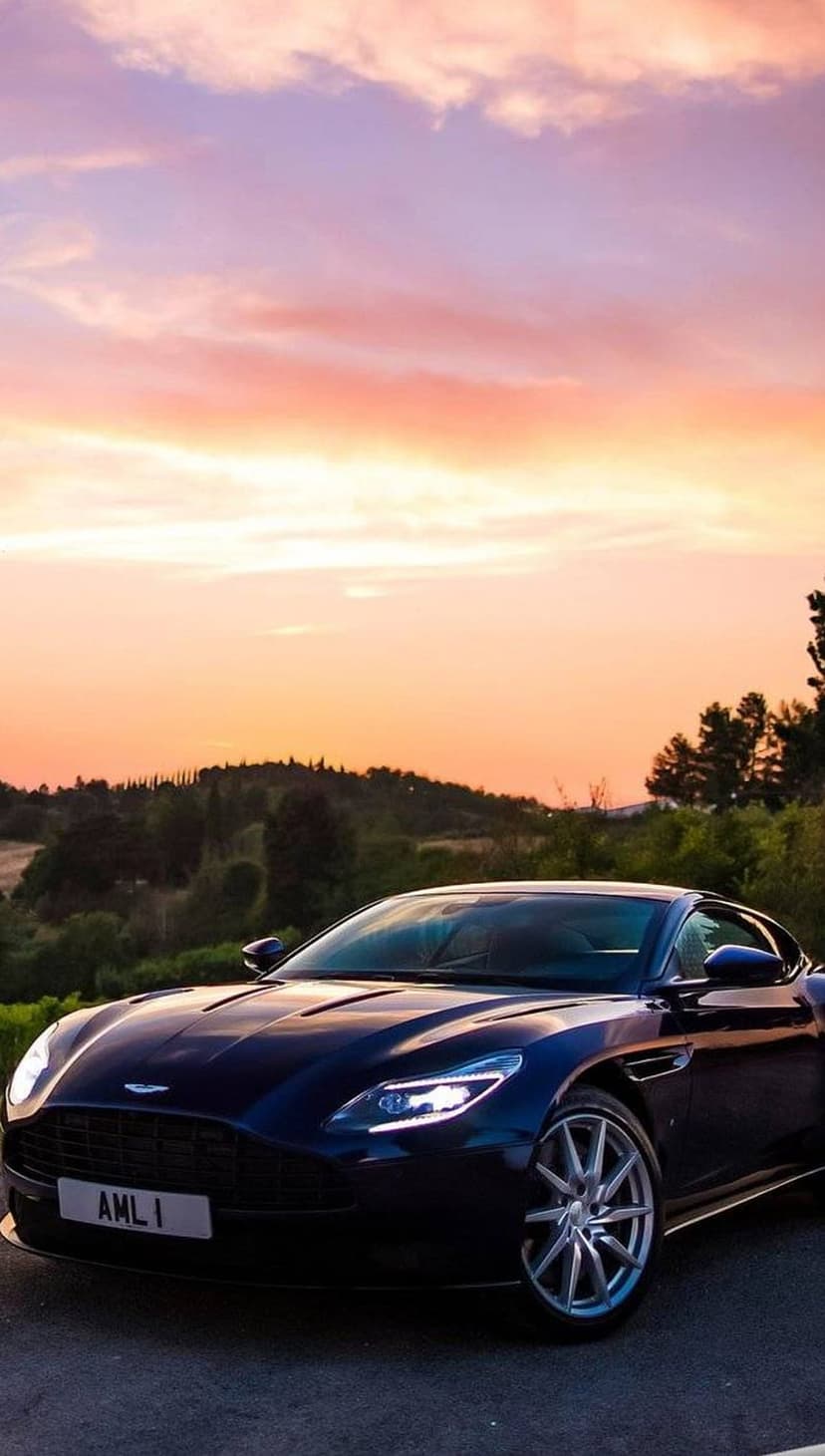 Aston Martin DB11 with glowing headlights on a Tuscan road during a vibrant orange and purple sunset.