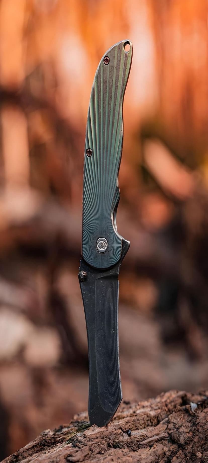 Tactical folding knife with a green sunburst handle and dark cleaver blade stuck into a textured wooden block.