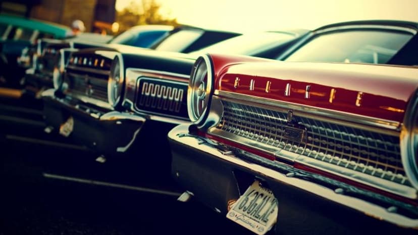 Close-up of a red vintage car's chrome grille and headlights at sunset with other classic cars blurred behind.