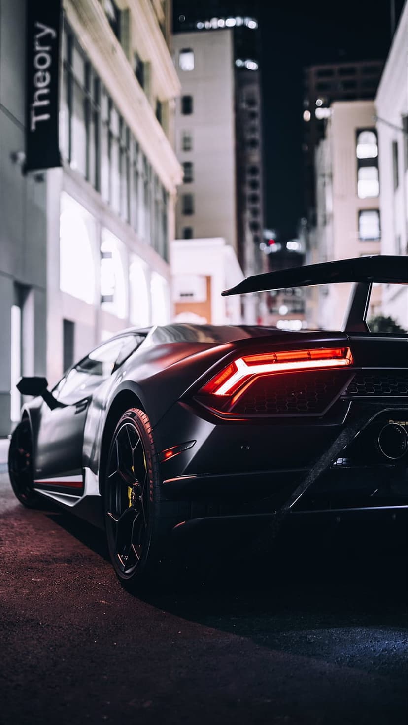 Black Lamborghini rear view on a dark street with glowing red taillights and neon reflections on the asphalt.