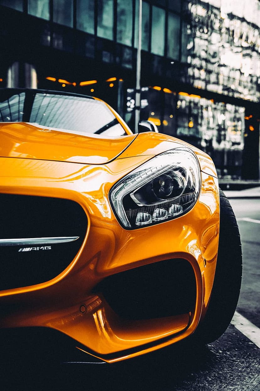 Orange Mercedes-AMG GT sports car headlight and radiator grille detail on a blurred urban city street background.