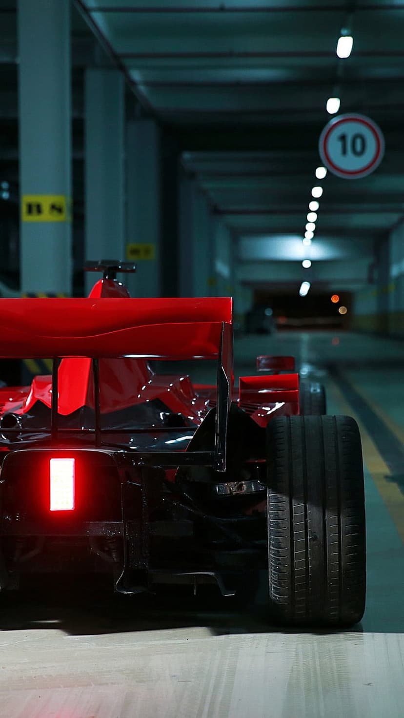 Red Formula 1 race car rear wing and glowing taillight in a dark concrete garage with soft overhead ceiling lights.