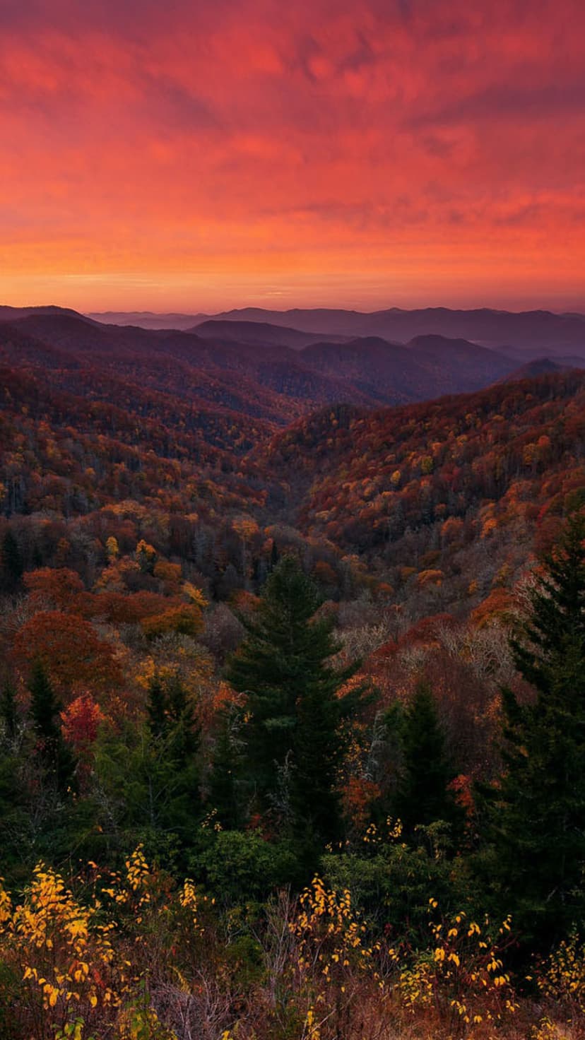 Rolling mountain ridges covered in orange fall trees sit beneath a vibrant red and orange atmospheric sunset sky.