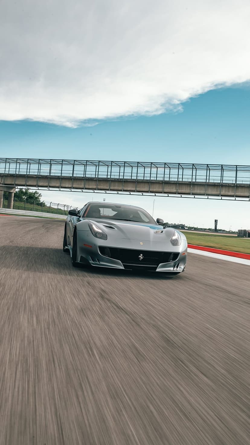 Silver Ferrari F12 TDF racing on a track with motion blur under a bridge featuring red and white curbs.