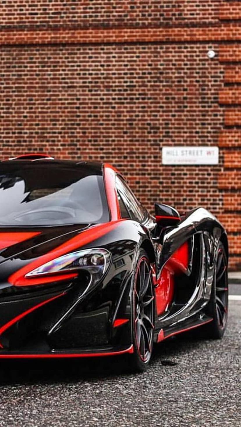 Black McLaren P1 hypercar with red accents parked on a cobblestone street by a brick wall and Hill Street sign.