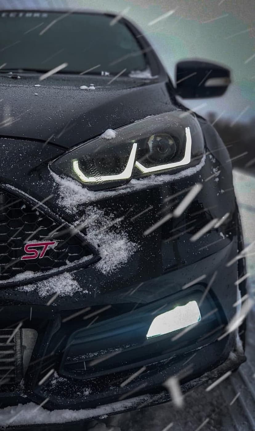 Ford Focus ST front grille and glowing LED headlights partially covered by falling snow in a dark winter scene.