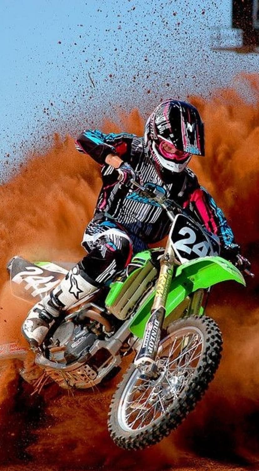 Motocross rider on a green bike jumps through an orange dust cloud against a blue sky mid-air during a race.